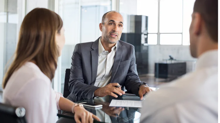 Businessman sits at table, pen in hand, with two business people