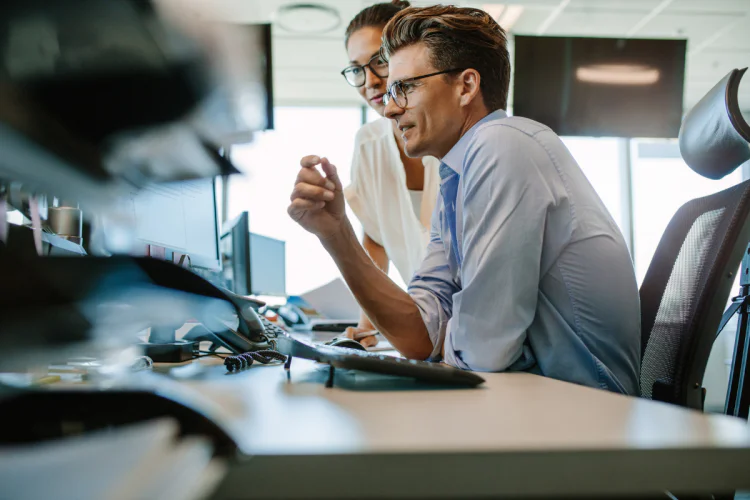 Business man sits at desk with female colleague standing beside him, both looking at computer