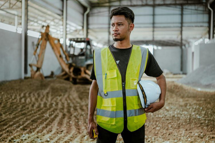 Worker in high-vis vest holds hard hat, excavator in background