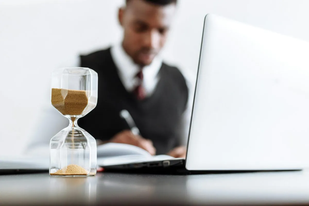 Business man works at laptop in background; hour glass sits in foreground