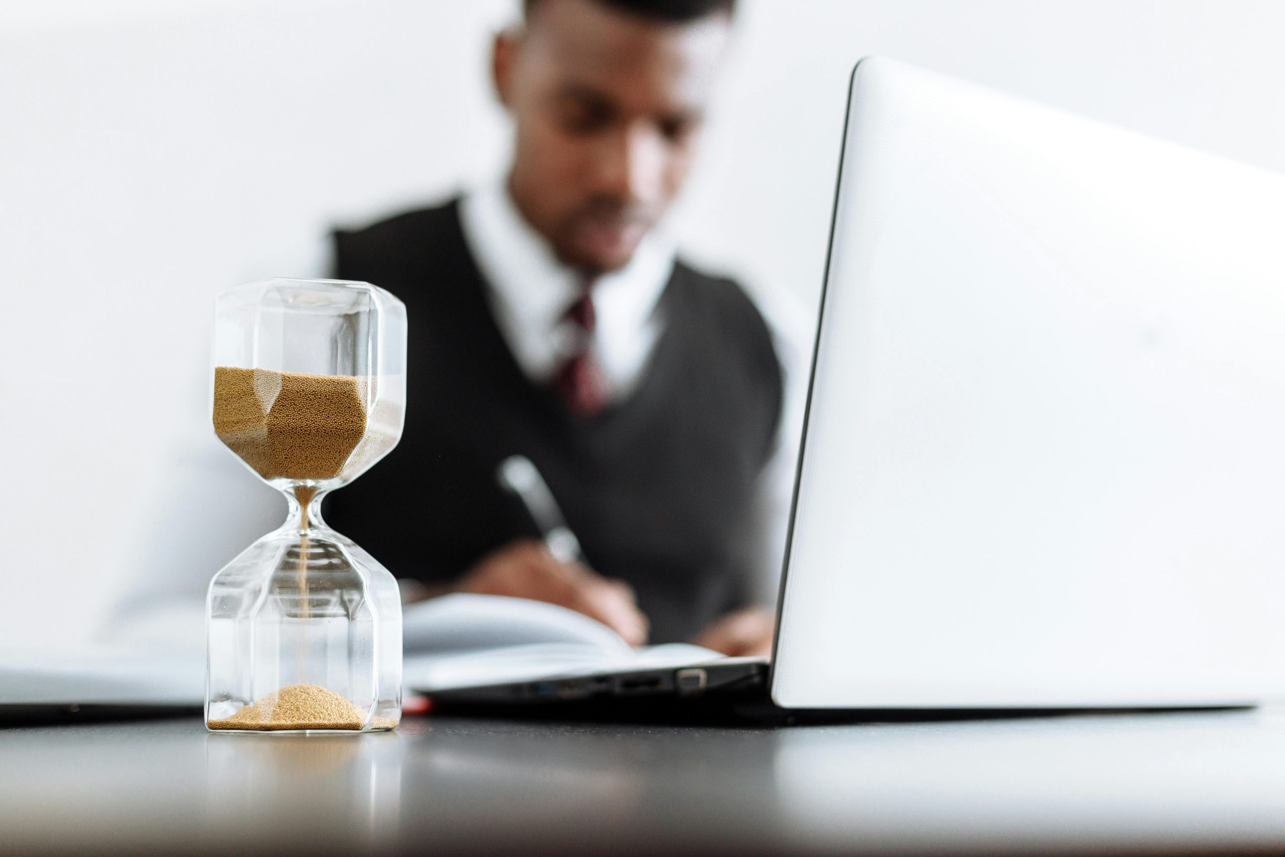 Business man works at laptop in background; hour glass sits in foreground