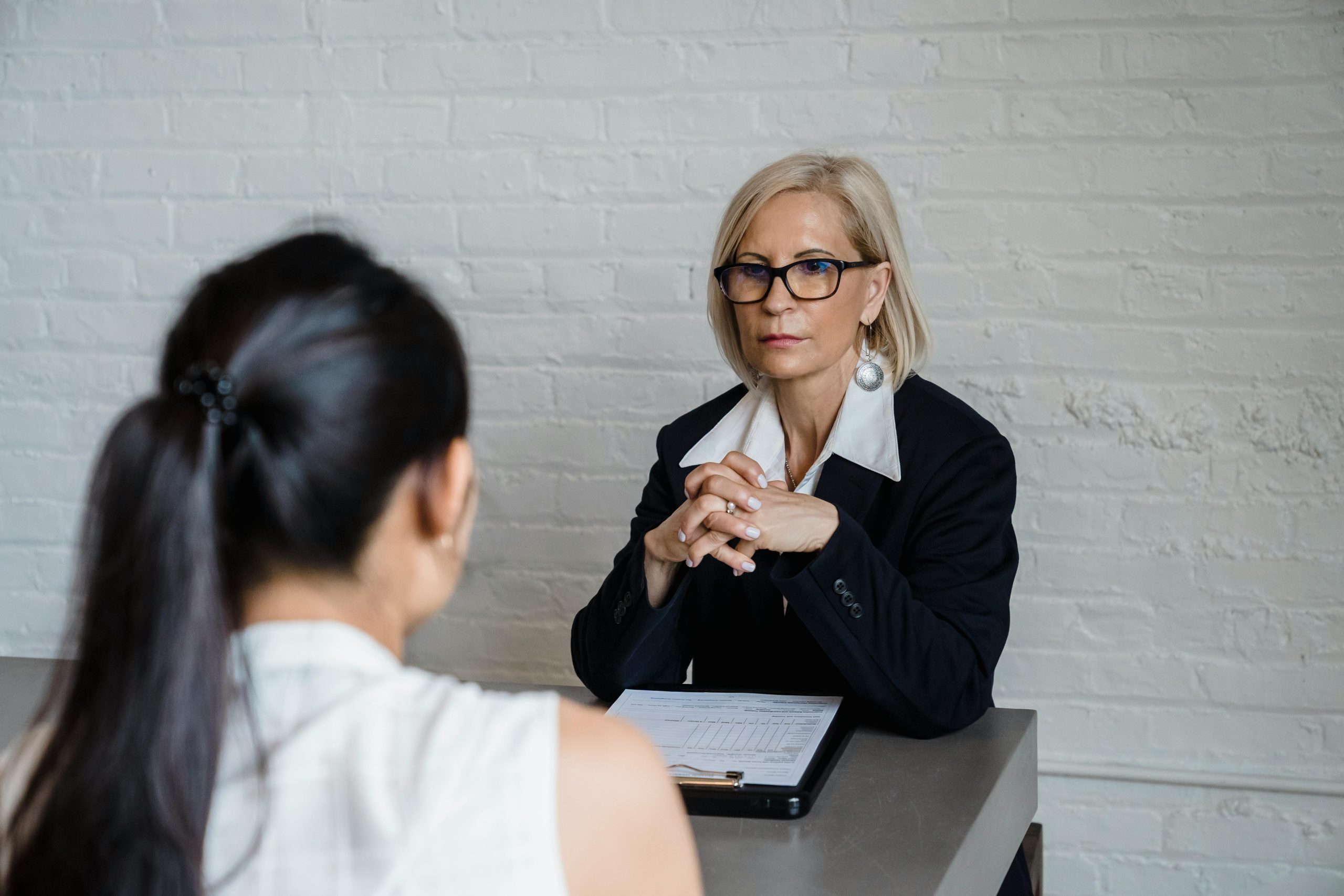 Blonde-haired business woman with serious expression sits across table from dark-haired business woman