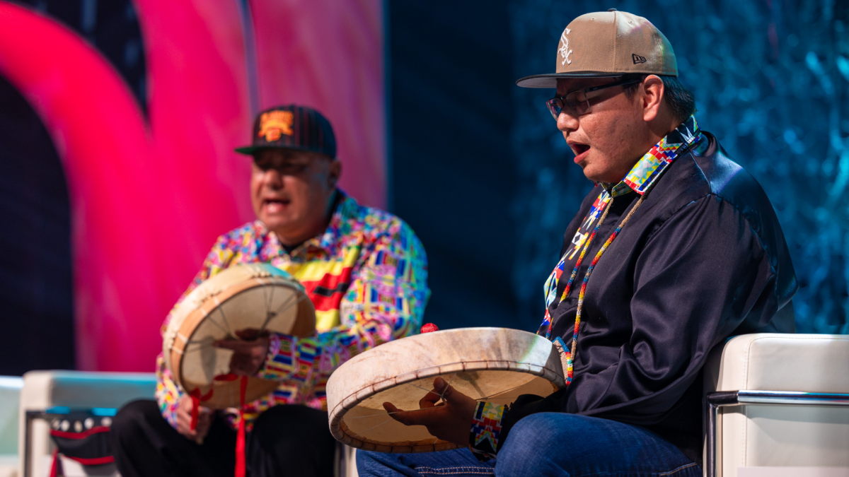 Two Indigenous men perform hand drumming while sitting on a stage.