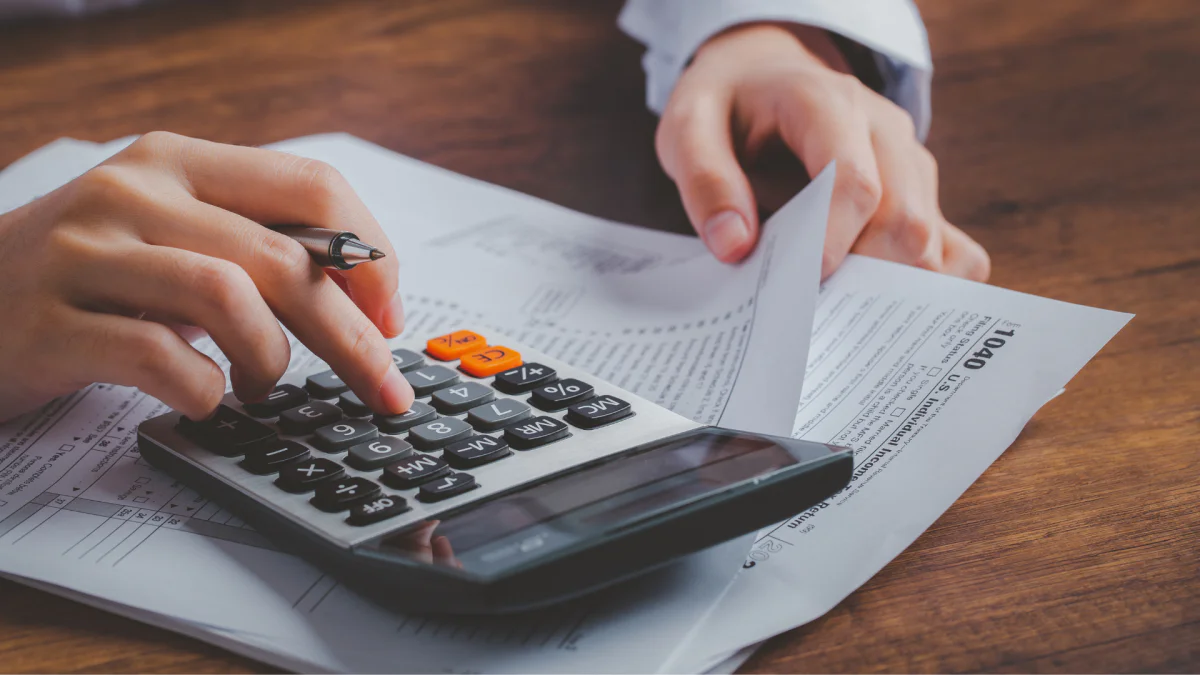 A close up photo of someone at a desk using a calculator and comparing to a stack of papers.