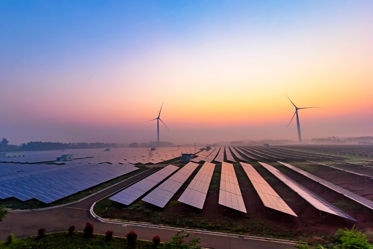 A sunset photo of a solar energy farm with windmills in the background