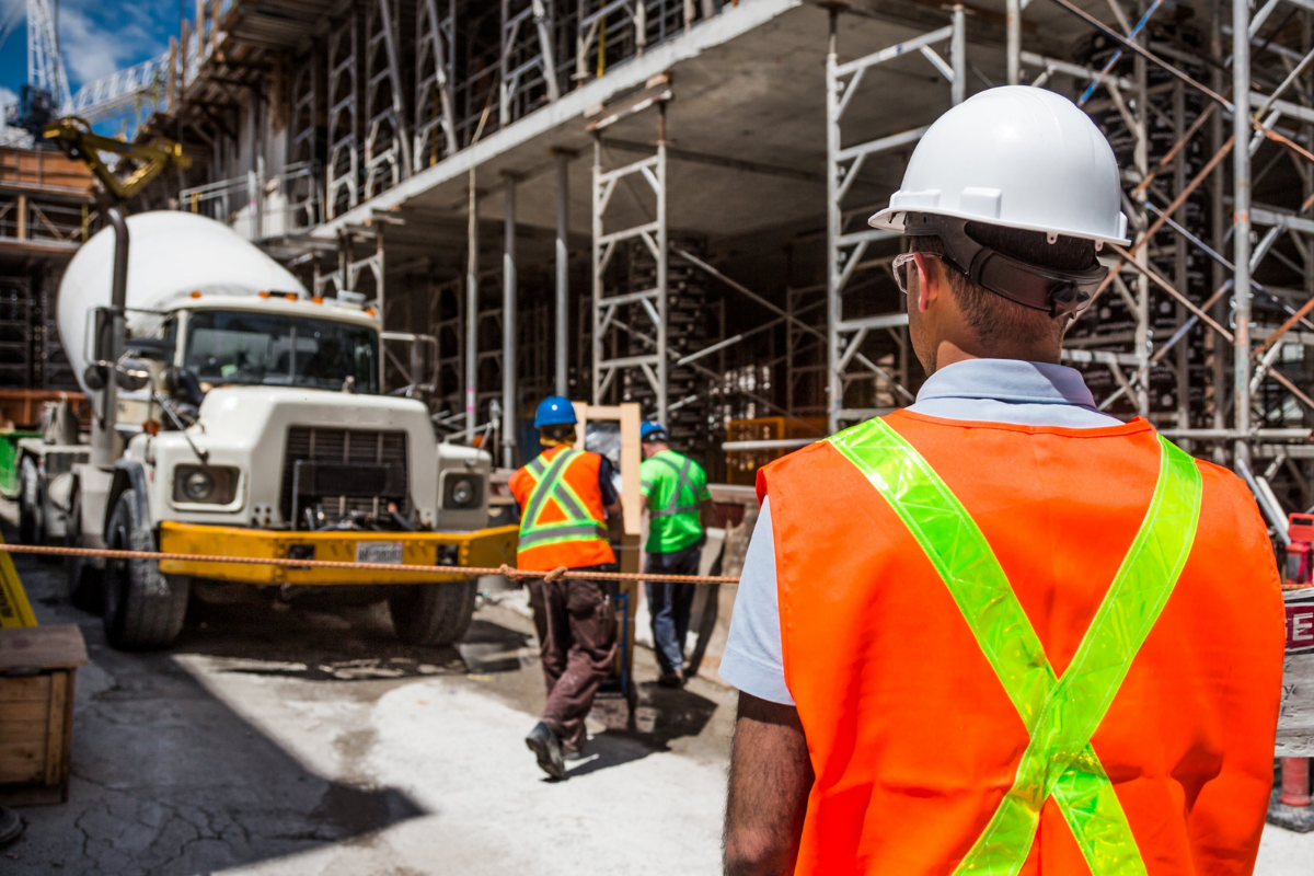 A construction site, with the focus on a worker standing with his back to the camera, a cement mixer in the background
