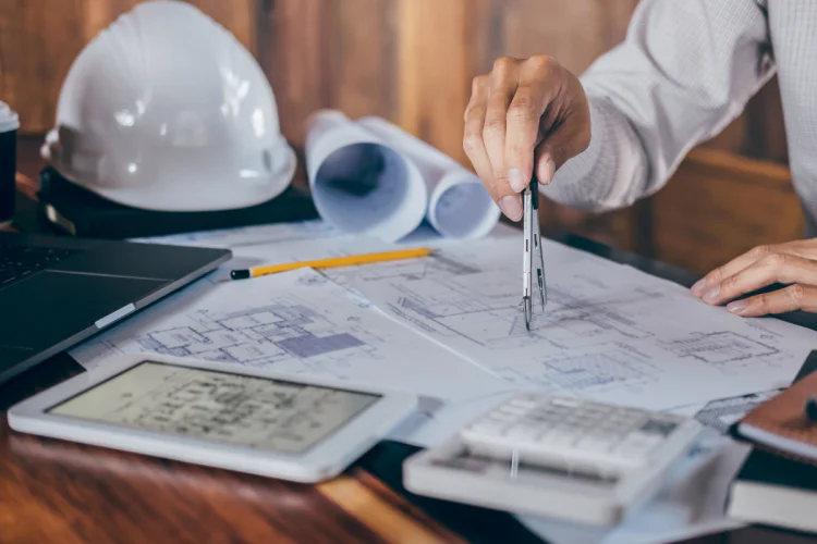 A close up photo of a drafting table with plans and a hard hat on it. An arm is reaching in from off screen.