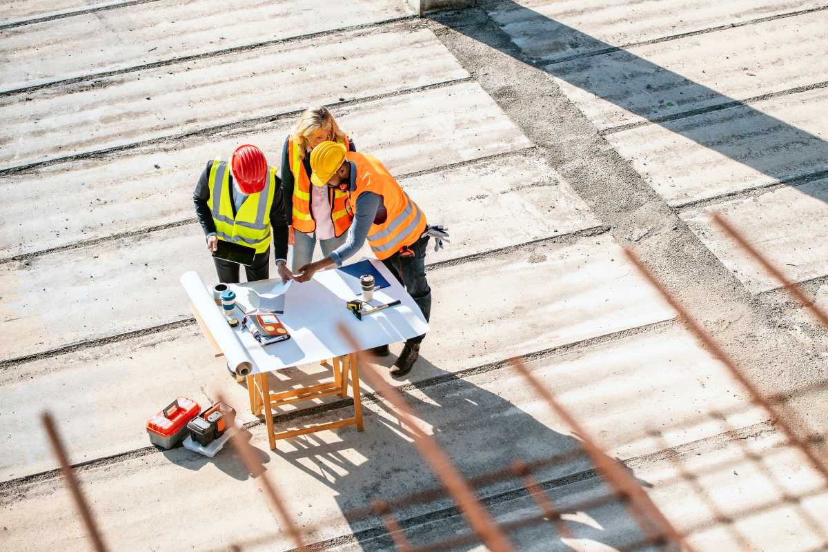 An overhead photo of three people in hard hats and vests looking over plans at a construction site