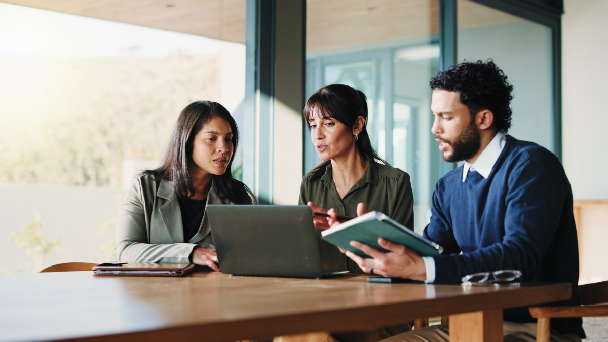 Three business people sit at table, looking at laptop