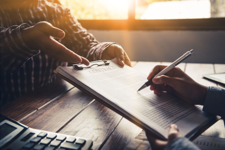 A close up photo of the signing of a contract over a desk. One party is passing the clipboard to another.