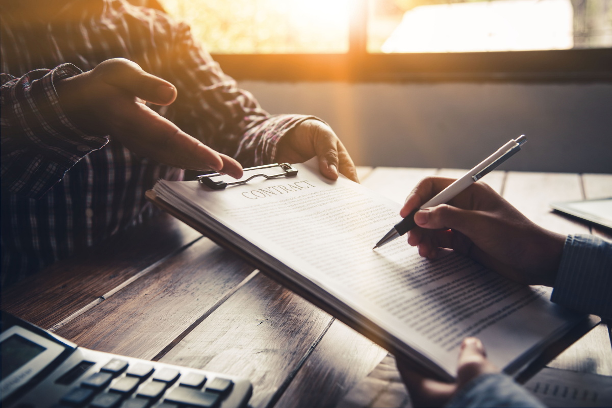 A close up photo of the signing of a contract over a desk. One party is passing the clipboard to another.