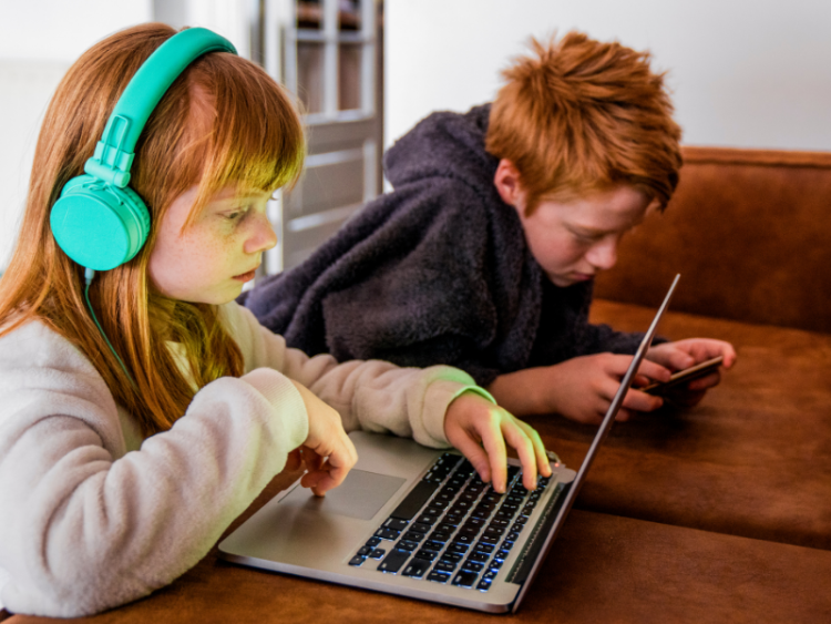 Two children at desk, one on a laptop with headphones on and one on a phone