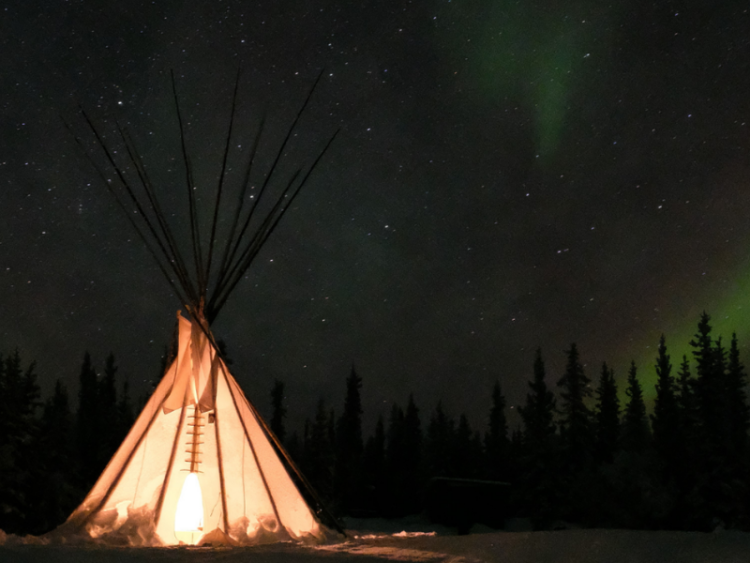 A teepee at night, lit from inside, with the northern lights shining overhead