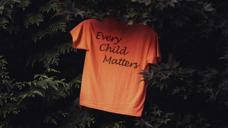 Orange tshirt hangs in greenery, text on shirt ready Every Child Matters
